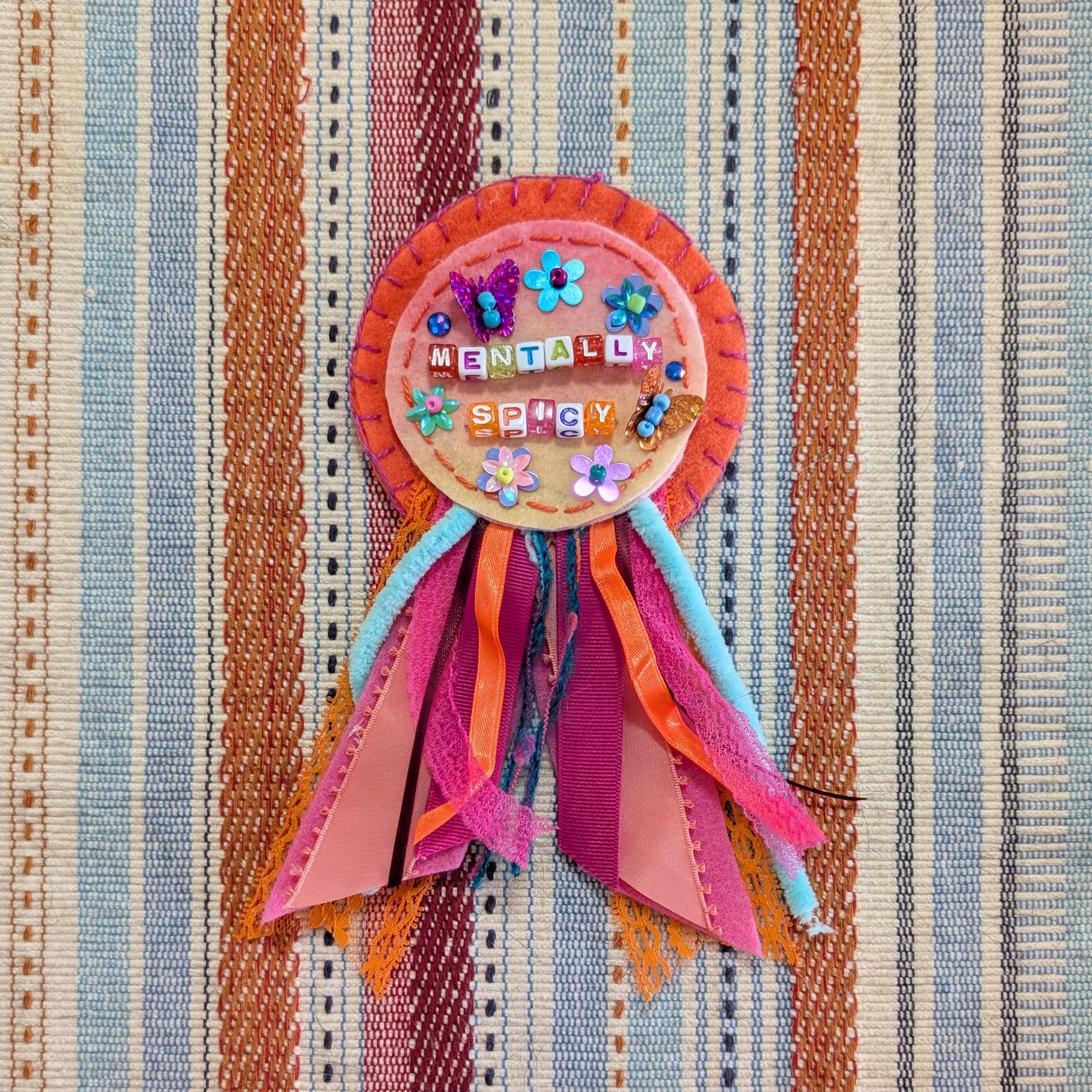 A colorful handmade ribbon award badge with a circular decorated center and multiple pleated paper ribbons, placed on a striped woven textile background.
