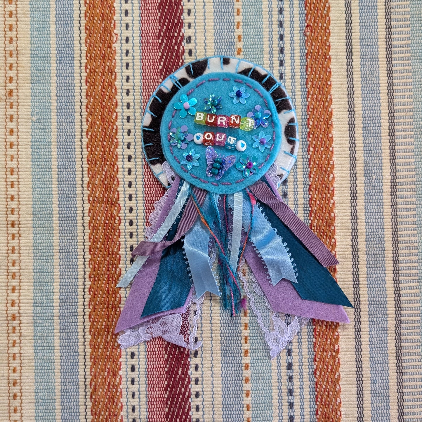 A handmade circular ribbon rosette with the words 'BURN IT OUT' in colorful letters, decorated with blue sequins and hanging ribbons in shades of blue, purple, and teal, laid flat on a striped woven textile background.