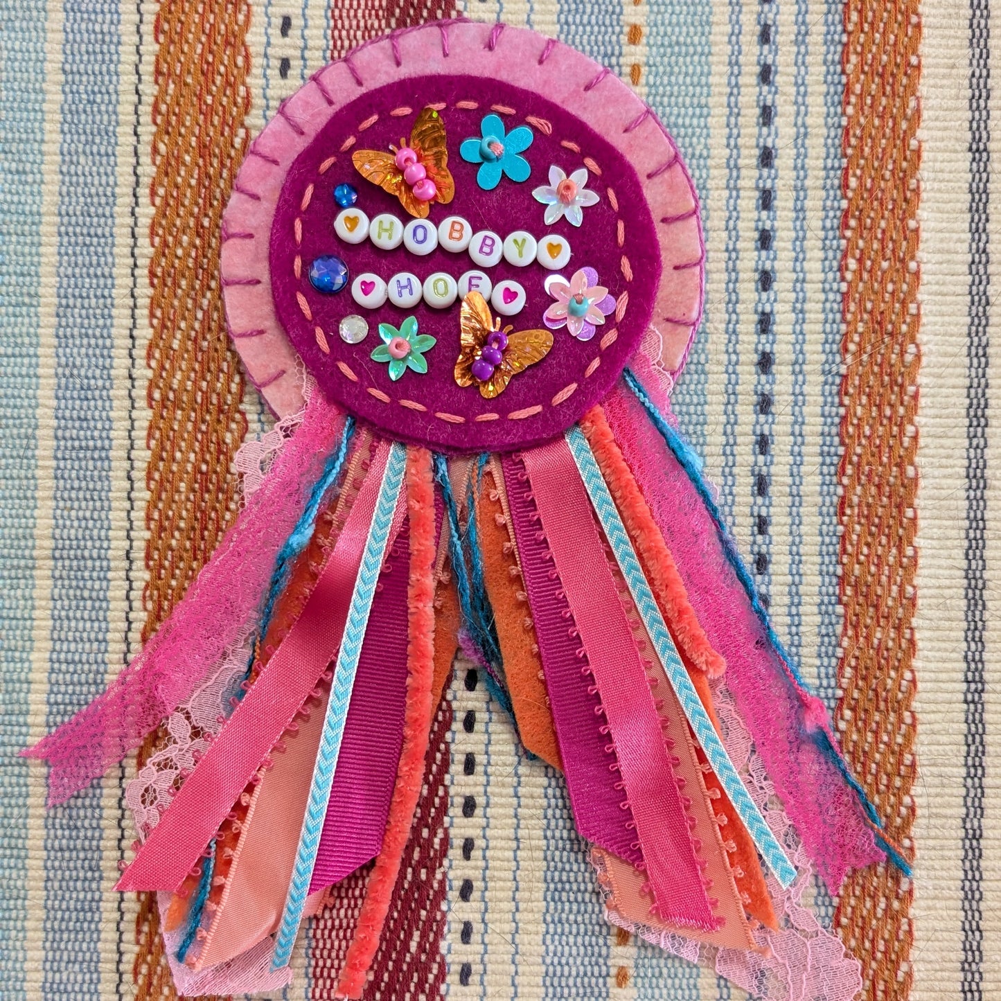 A colorful handmade ribbon brooch with a circular pink and purple center decorated with sequins and flowers, attached to a multi-colored fabric ribbon tail, displayed on a striped woven textile background.
