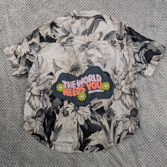 A short-sleeve button-up shirt with a black and white floral print and a colorful graphic patch reading 'THE WORLD NEEDS YOU' laid flat on a gray textured blanket.
