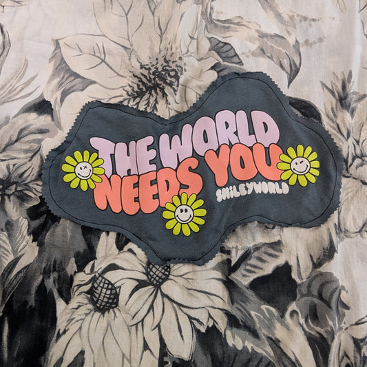 A close-up of a black cloud-shaped patch with the text 'THE WORLD NEEDS YOU' in pink and orange, decorated with yellow flowers, placed on a black and white floral-patterned garment.