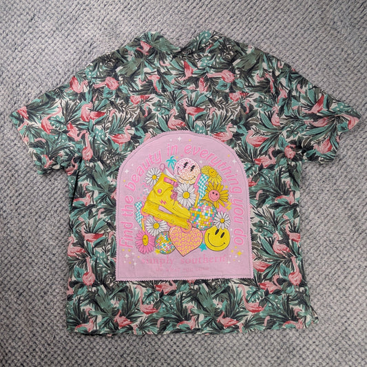 A folded garment laid flat on a textured gray carpet, featuring a pink tropical leaf print with green and pink tones and a large pink semicircular patch with a colorful cartoon graphic.