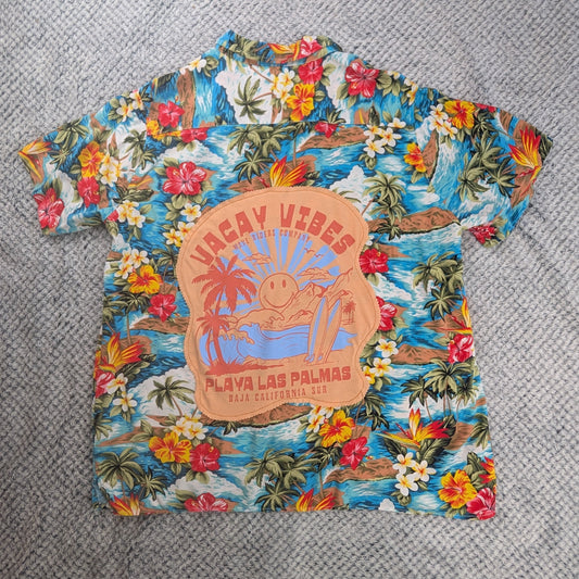 A short-sleeve Hawaiian shirt with a colorful tropical floral print and a central graphic reading 'Canyans Beach Playa Las Palmas', laid flat on a textured gray blanket with a person's feet visible at the bottom edge.