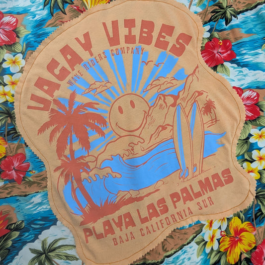 A close-up of a tropical beach-themed fabric featuring a large tan patch with 'Vacay Vibes' and 'Playa Las Palmas' text, surrounded by blue waves, palm trees, and colorful flowers.