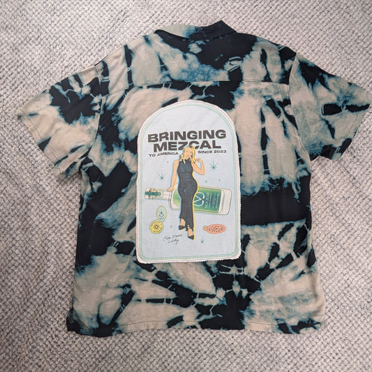 A tie-dye t-shirt with a black, white, and blue pattern, featuring a white graphic patch on the front that reads 'BRINGING THE MEZCAL'. The shirt is laid flat on a textured beige rug.