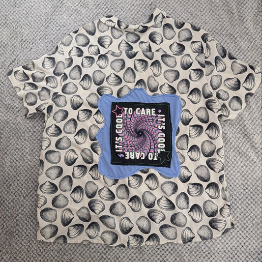 A short-sleeve t-shirt with a black and white seashell pattern, laid flat on a gray knit blanket. The center features a graphic patch with a purple spiral and the text 'TO CARE IT'S GOOD TO CARE'.