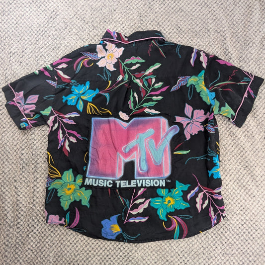 A black short-sleeve button-up shirt with a colorful floral print and a large MTV Music Television logo on the front, laid flat on a textured white blanket with a person's feet visible at the bottom edge.