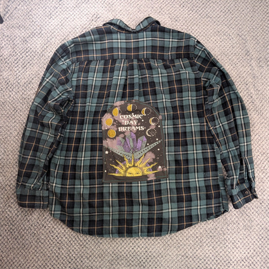 A green and black plaid long-sleeve button-up shirt laid flat on a textured white blanket, featuring a colorful graphic print on the back with the words 'Sugared Love Affair' and 'Love Never Dies'.