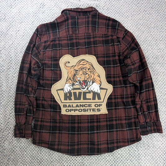 A red and black plaid flannel shirt jacket laid flat on a textured white rug, featuring a large graphic patch on the back with a tiger and the text 'RVCA BALANCE OF OPPOSITES'.
