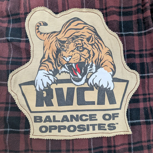 A close-up of a red and black plaid shirt featuring a large embroidered patch with a tiger graphic and the text 'RVCA' and 'BALANCE OF OPPOSITES'.