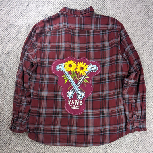 A red and black plaid flannel shirt laid flat on a white textured blanket, featuring a large back graphic with crossed bones, yellow sunflowers, and the text 'VANS' and 'EST. 2015'.