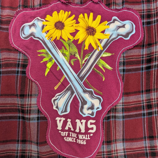 A close-up of a red and black plaid fabric surface with a pink embroidered patch featuring crossed bones, yellow flowers, green leaves, and the text 'VANS OFF THE WALL SINCE 1966'.