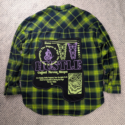A green and black plaid flannel shirt with a large graphic patch on the back, laid flat on a textured beige carpet. The patch features the text 'HUSTLE' and purple and green graphics.