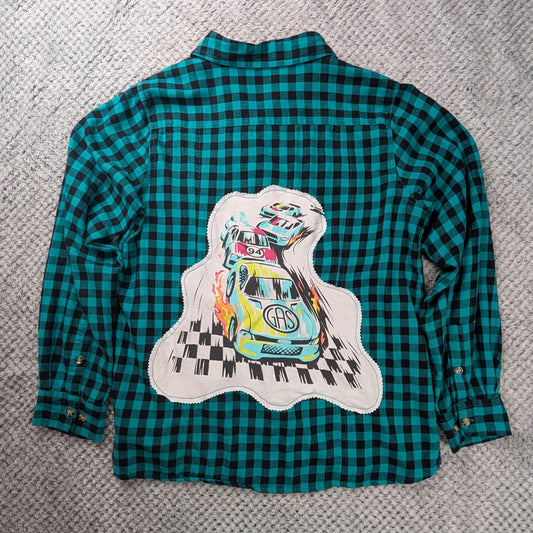 A teal and black checkered long-sleeve flannel shirt laid flat on a gray textured blanket, featuring a large graphic print of a cartoon car on a white cloud shape on the back.