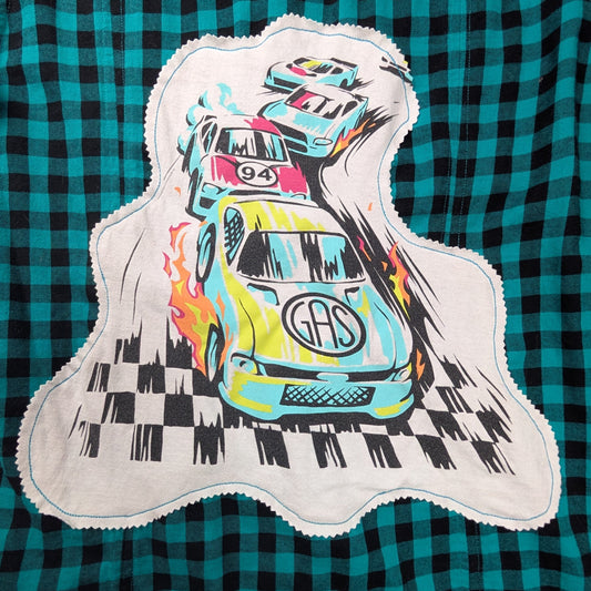 A close-up of a green and black plaid fabric item, likely a shirt or blanket, featuring a large applique patch with a colorful cartoon race car graphic.