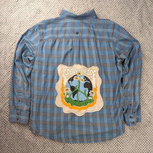 A blue and gray plaid long-sleeve button-up shirt laid flat on a textured grey blanket, featuring a large 'Grow Peace' patch on the back.