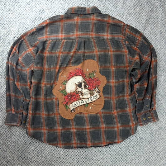 A long-sleeve plaid flannel shirt laid flat on a gray textured rug. The shirt features a brown and orange plaid pattern with a large graphic patch on the back depicting a skull, roses, and a banner that reads 'WILD & FREE'.