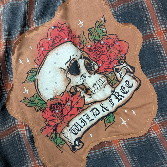 A close-up of a plaid fabric garment, likely a shirt or jacket, featuring a large tan patch with a skull, red flowers, green leaves, and a banner reading 'WILD FREE'.