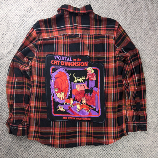 A red and black plaid flannel shirt with a colorful purple and orange graphic patch, laid flat on a gray carpet. graphic says 'portal to the cat dimension'. 