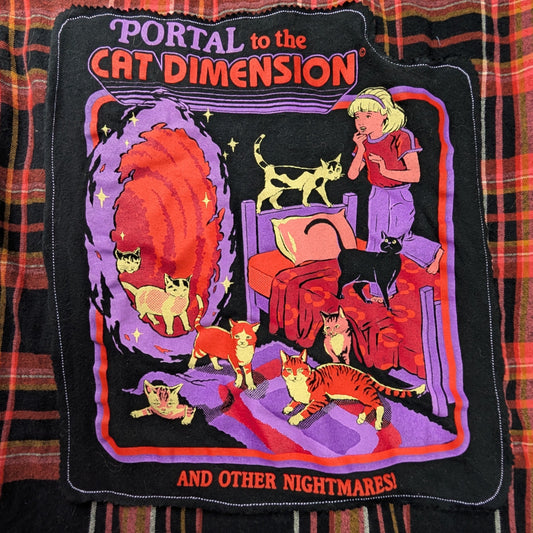 A red and black plaid flannel shirt with a colorful purple and orange graphic patch, laid flat on a gray carpet. graphic says 'portal to the cat dimension'. 