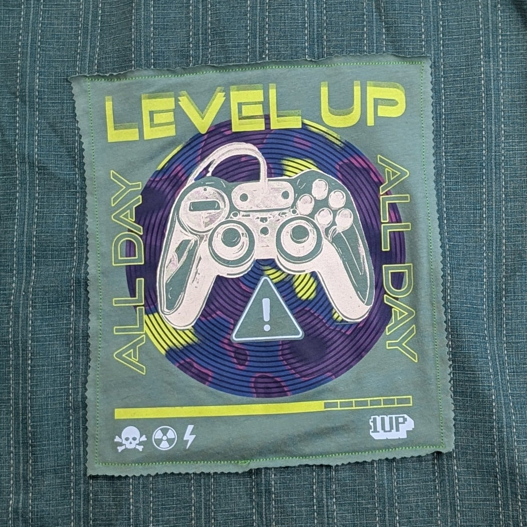 A dark teal-green short-sleeve button-up shirt with vertical pinstripes, featuring a colorful retro video game 'Level Up' graphic patch on the back, laid flat on a textured gray blanket.