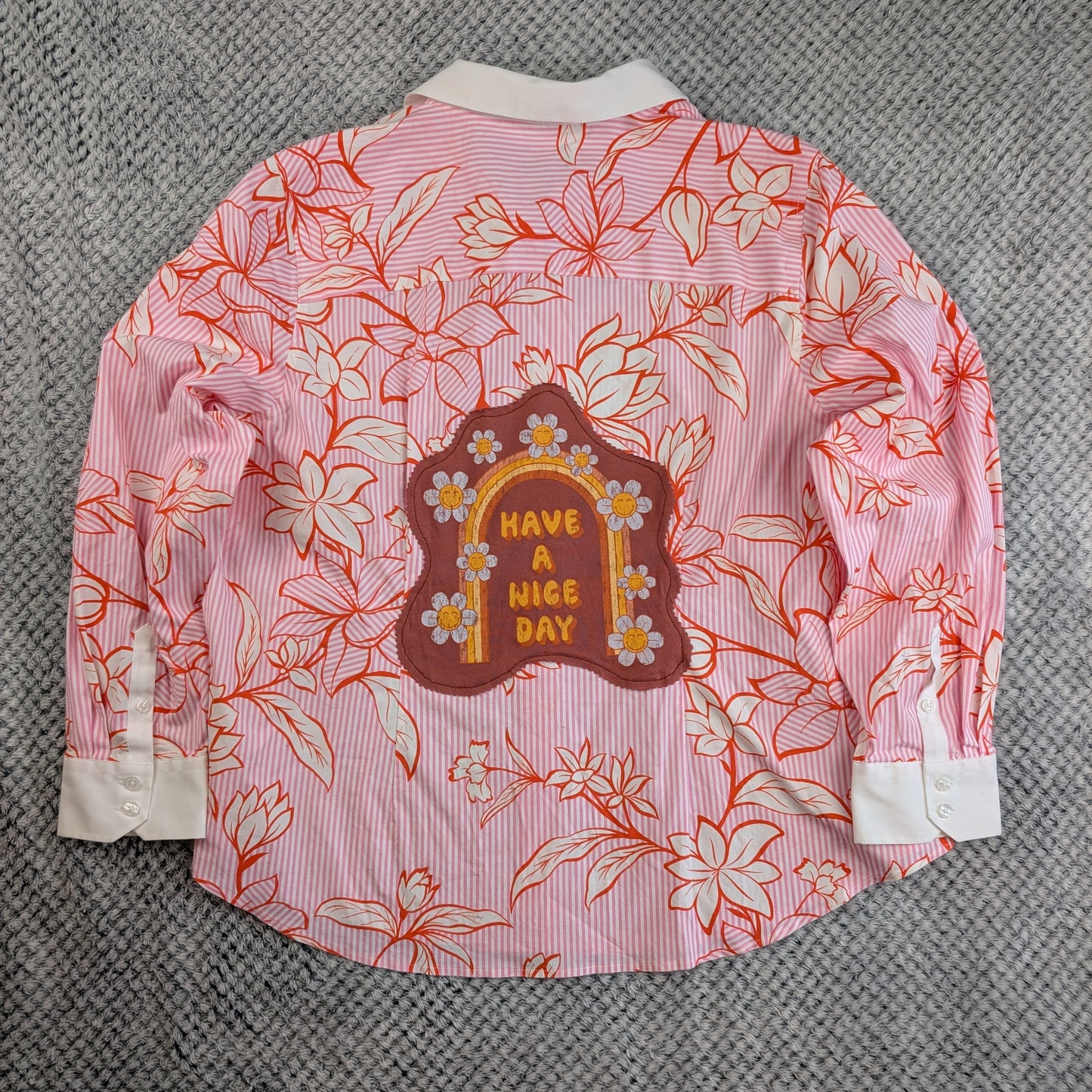 A pink and white striped long-sleeve button-up shirt with a red-orange floral line-art print, featuring a brown arch-shaped patch on the back with yellow text reading 'HAVE A NICE DAY' and small white flowers. The shirt is laid flat on a gray textured fabric background.