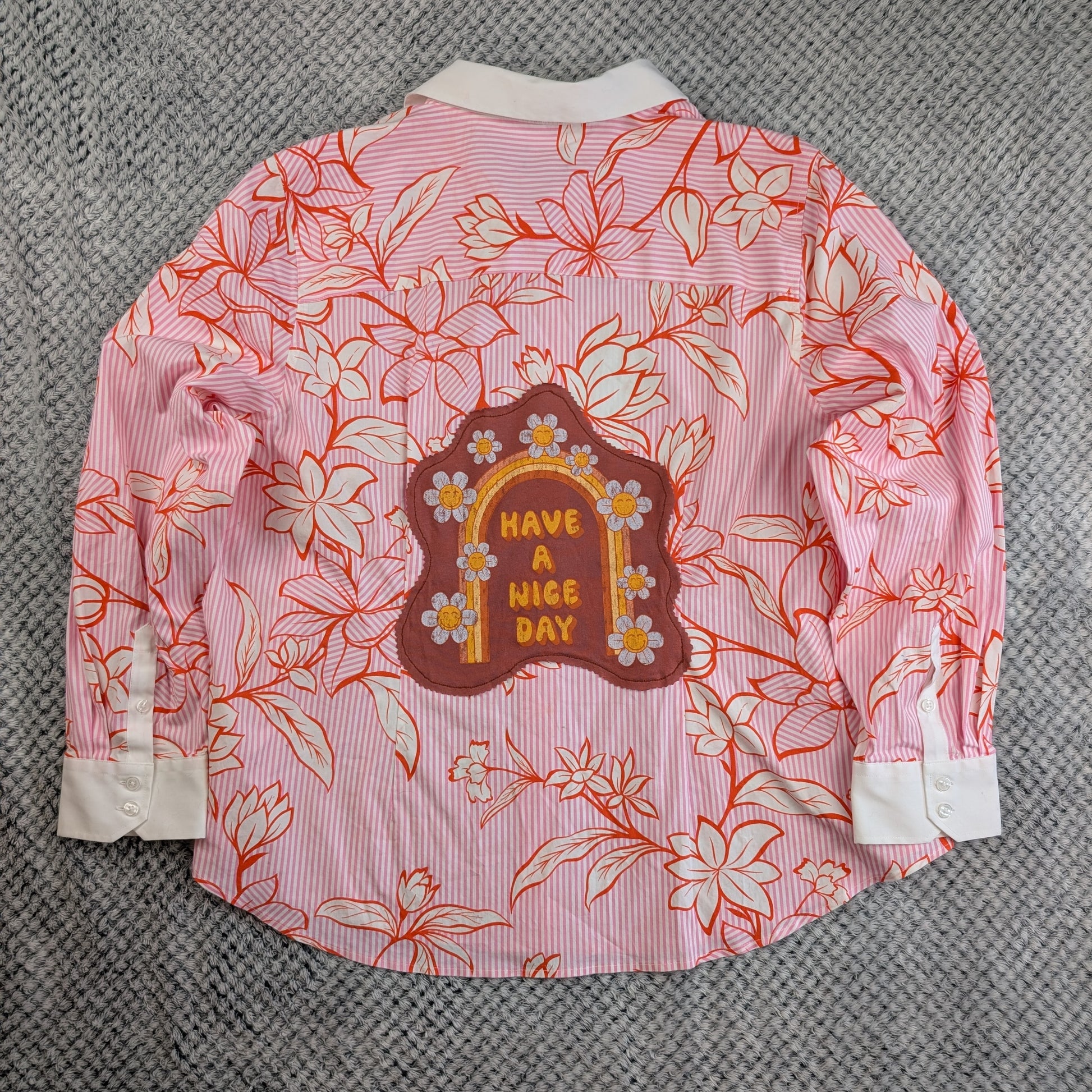 A pink and white striped long-sleeve button-up shirt with a red-orange floral line-art print, featuring a brown arch-shaped patch on the back with yellow text reading 'HAVE A NICE DAY' and small white flowers. The shirt is laid flat on a gray textured fabric background.