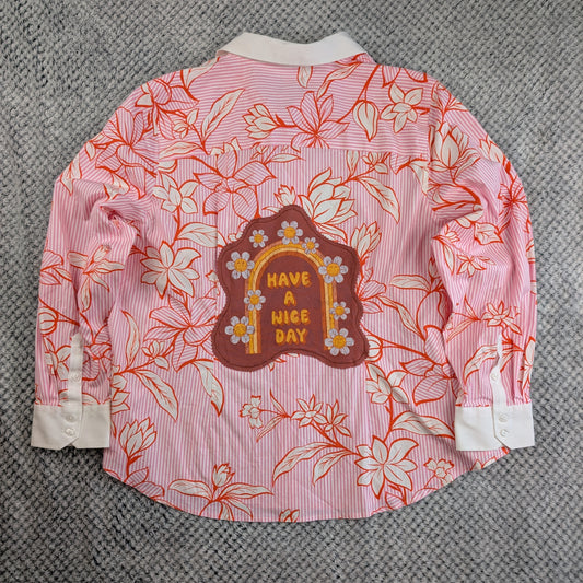 A pink and white striped long-sleeve button-up shirt with a red-orange floral line-art print, featuring a brown arch-shaped patch on the back with yellow text reading 'HAVE A NICE DAY' and small white flowers. The shirt is laid flat on a gray textured fabric background.
