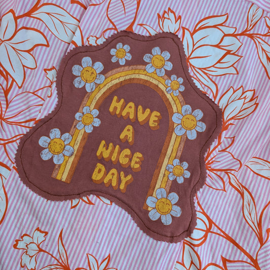 A close-up of a decorative fabric patch with scalloped edges, featuring a brown background, yellow embroidered arch and text reading 'HAVE A NICE DAY,' and blue-and-white daisy flowers. The patch is placed on pink striped fabric with orange floral outlines.