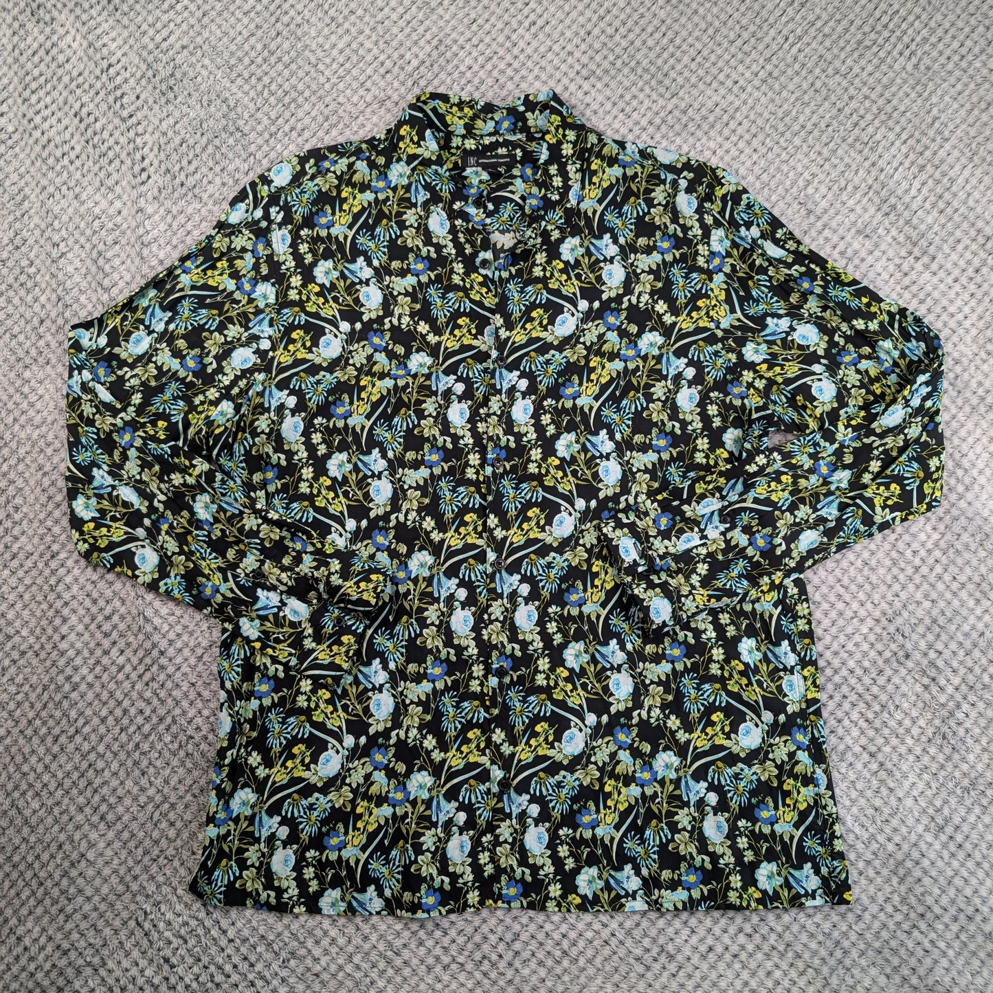 A long-sleeve button-up shirt with a dense floral and bird pattern in blue, yellow, and white on a black background, laid flat on a gray textured knit surface.