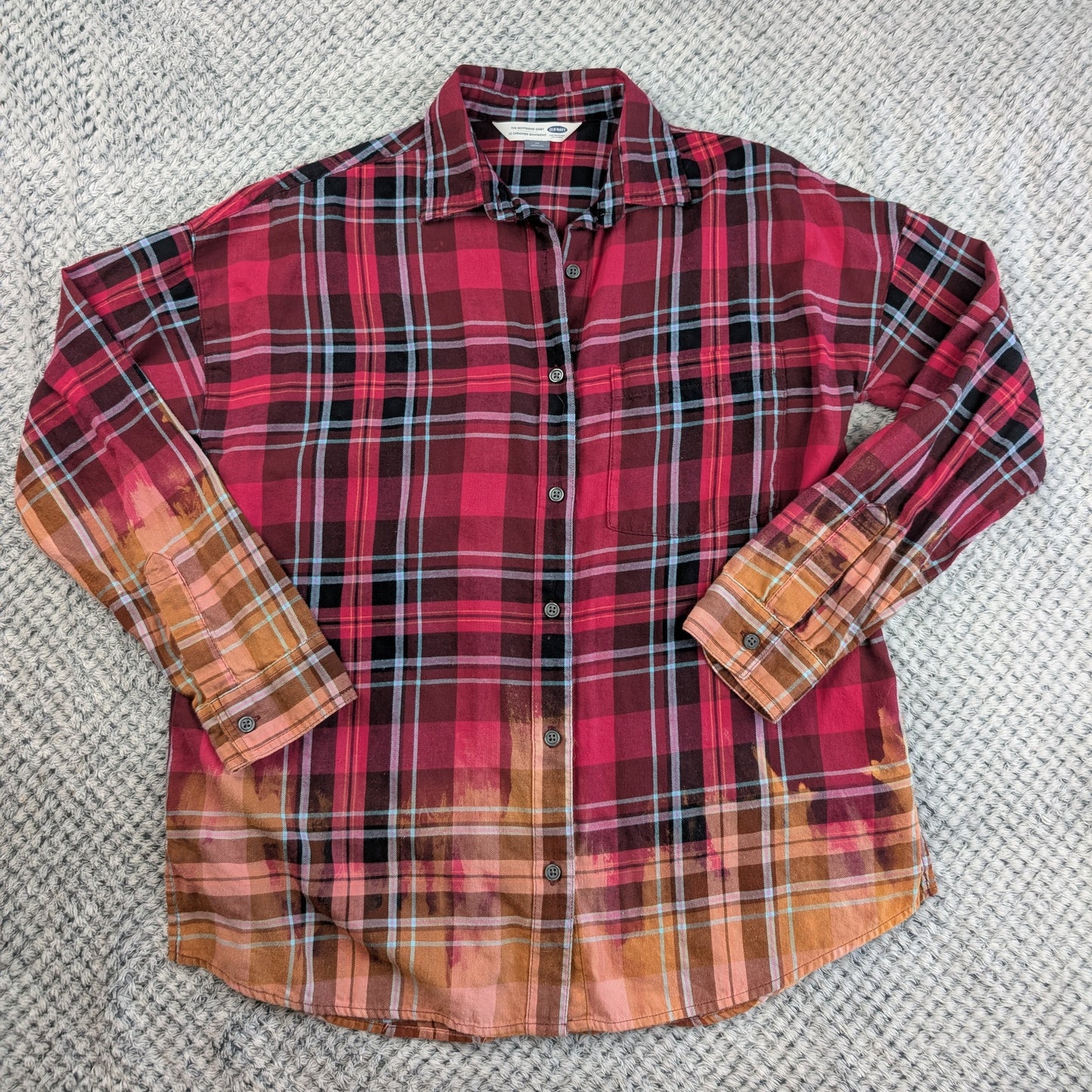 A long-sleeve button-up plaid shirt laid flat on a textured gray carpet. The shirt features a red, black, and white plaid pattern on the upper body, fading into a warm orange and brown plaid pattern on the lower hem and sleeves.