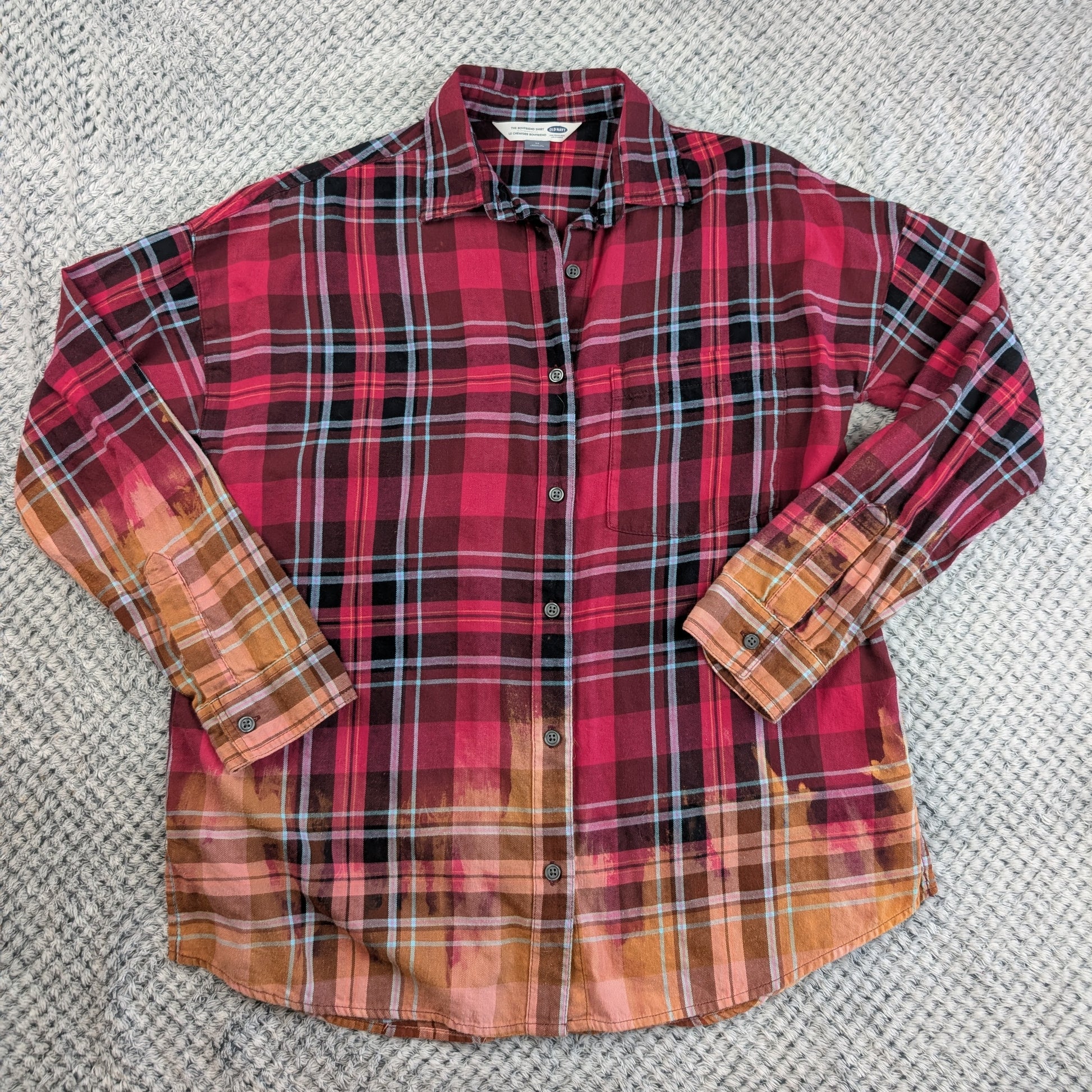 A long-sleeve button-up plaid shirt laid flat on a textured gray carpet. The shirt features a red, black, and white plaid pattern on the upper body, fading into a warm orange and brown plaid pattern on the lower hem and sleeves.