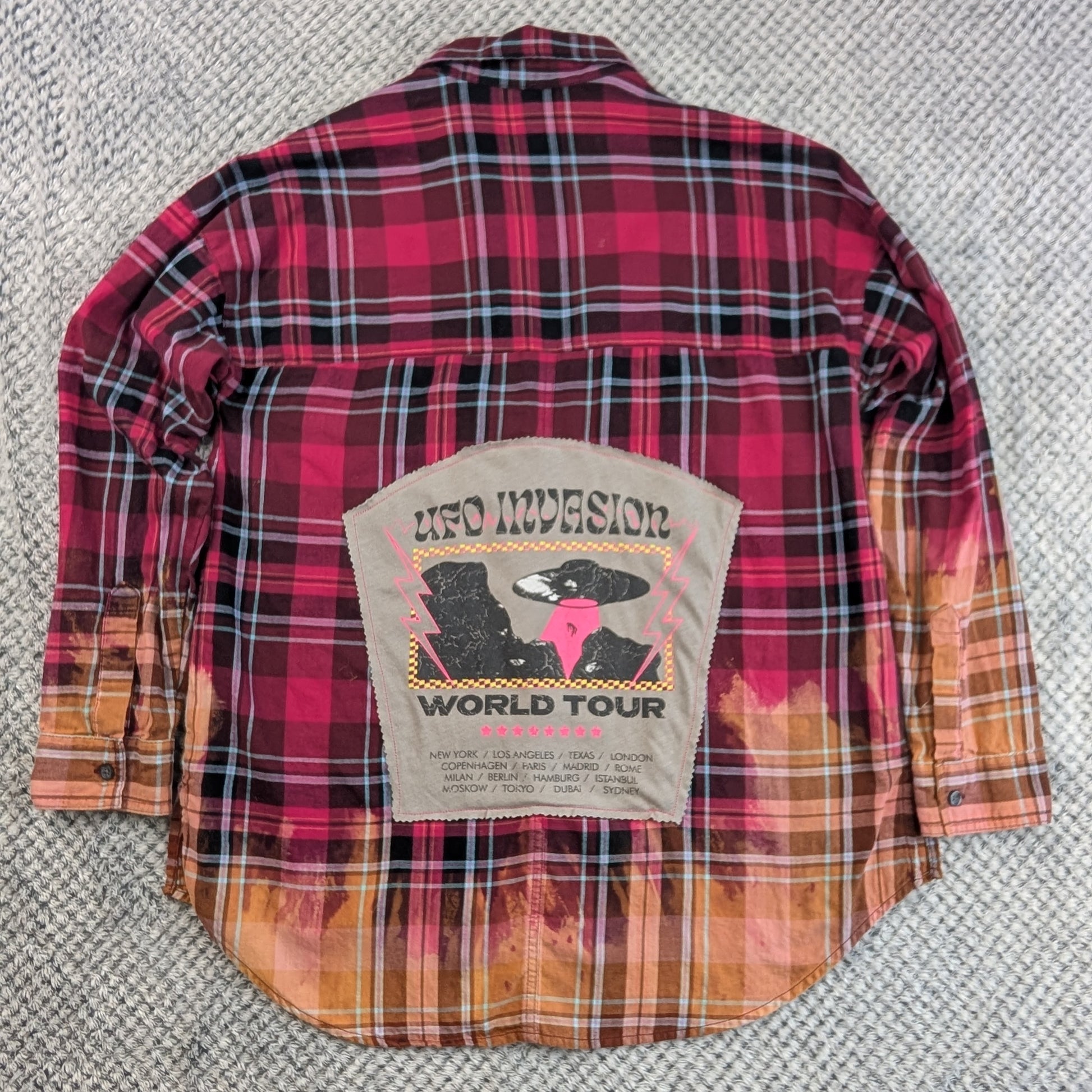A vintage red and black plaid flannel shirt jacket with orange and yellow gradient hem and sleeve cuffs, featuring a large 'UFO Invasion World Tour' patch on the back, laid flat on a gray textured carpet.