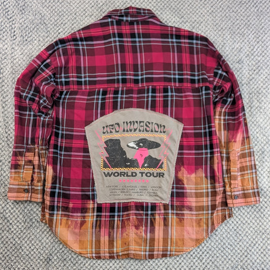 A vintage red and black plaid flannel shirt jacket with orange and yellow gradient hem and sleeve cuffs, featuring a large 'UFO Invasion World Tour' patch on the back, laid flat on a gray textured carpet.
