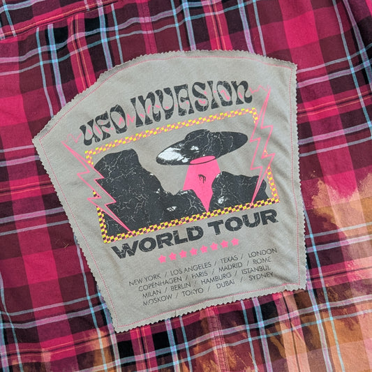 A close-up of a vintage band merchandise item featuring a beige embroidered patch for 'UFO Impression World Tour' with a raven graphic and pink accents, laid flat on a red and pink plaid flannel shirt.