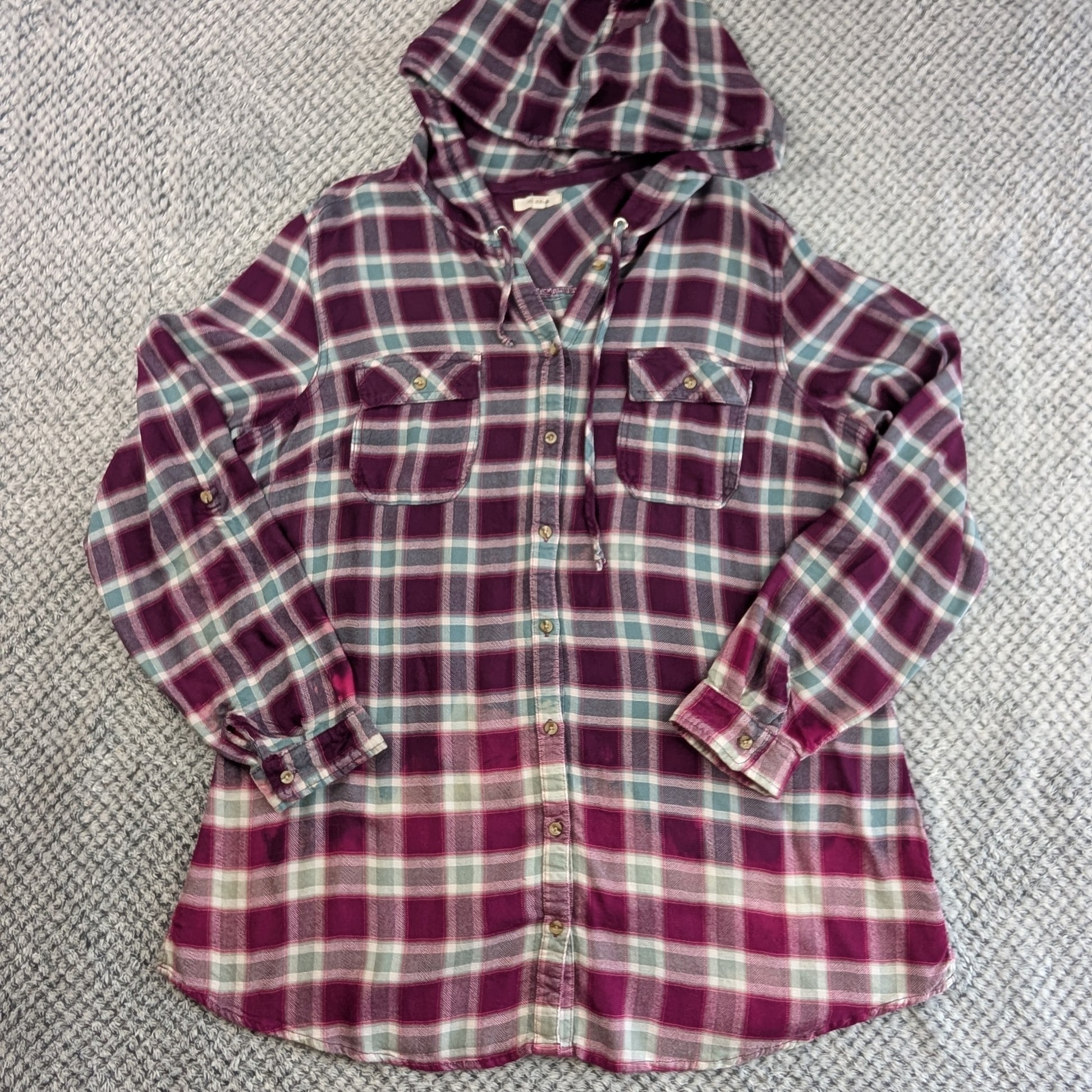 A long-sleeve hooded button-up flannel shirt in a purple, teal, and white plaid pattern, laid flat on a gray textured carpet.