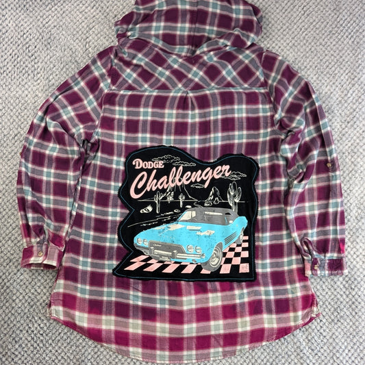 A long-sleeve plaid flannel shirt laid flat on a textured gray carpet. The shirt features a magenta, purple, and white plaid pattern with a large graphic patch on the front depicting a blue classic car and the text 'Dodge Challenger'.