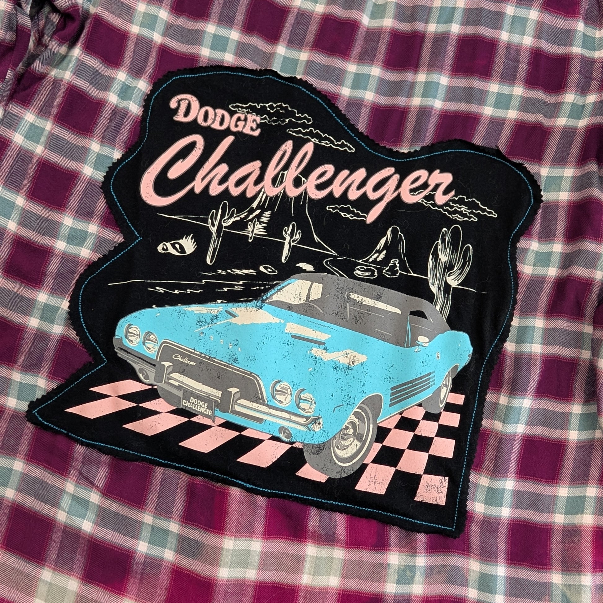 A distressed embroidered patch featuring a light blue Dodge Challenger car and the words 'Dodge Challenger' in pink, placed on a purple and green plaid fabric background.