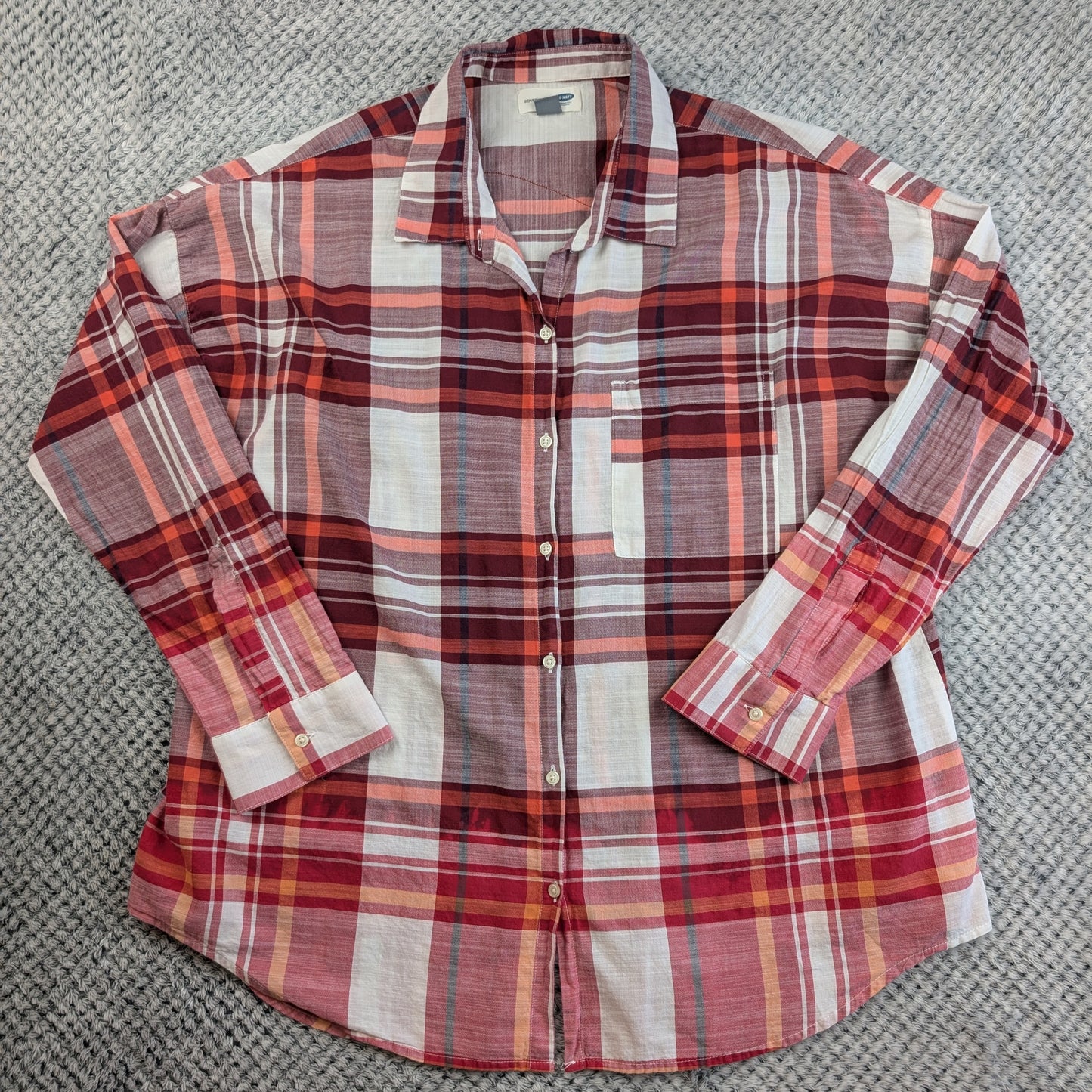 A long-sleeve plaid button-up shirt laid flat on a textured gray carpet. The shirt features a red, orange, and white plaid pattern with thin blue and green accent lines.