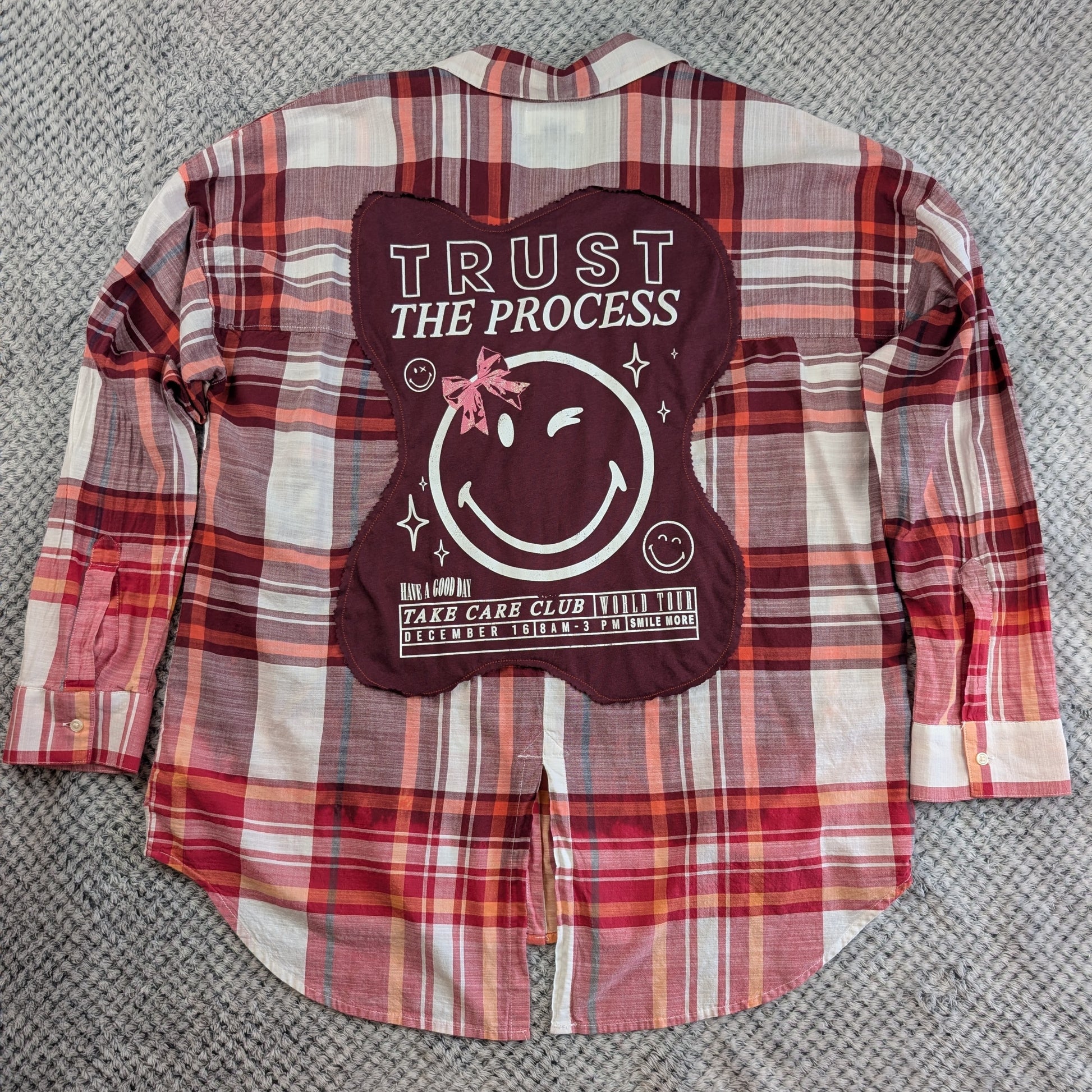 A red, pink, and white plaid flannel shirt laid flat on a gray textured carpet, featuring a large dark red back graphic with a smiley face, stars, and the text 'TRUST THE PROCESS' and 'TAKE CARE CLUB'.