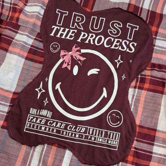 A maroon bone-shaped fabric patch with a white smiley face graphic, pink bow detail, and printed text reading 'TRUST THE PROCESS' and 'HAVE A GOOD DAY TAKE CARE CLUB WILD PARTY DECEMBER 16 8AM-3 PM SMILE MORE', laid flat on a plaid blanket.