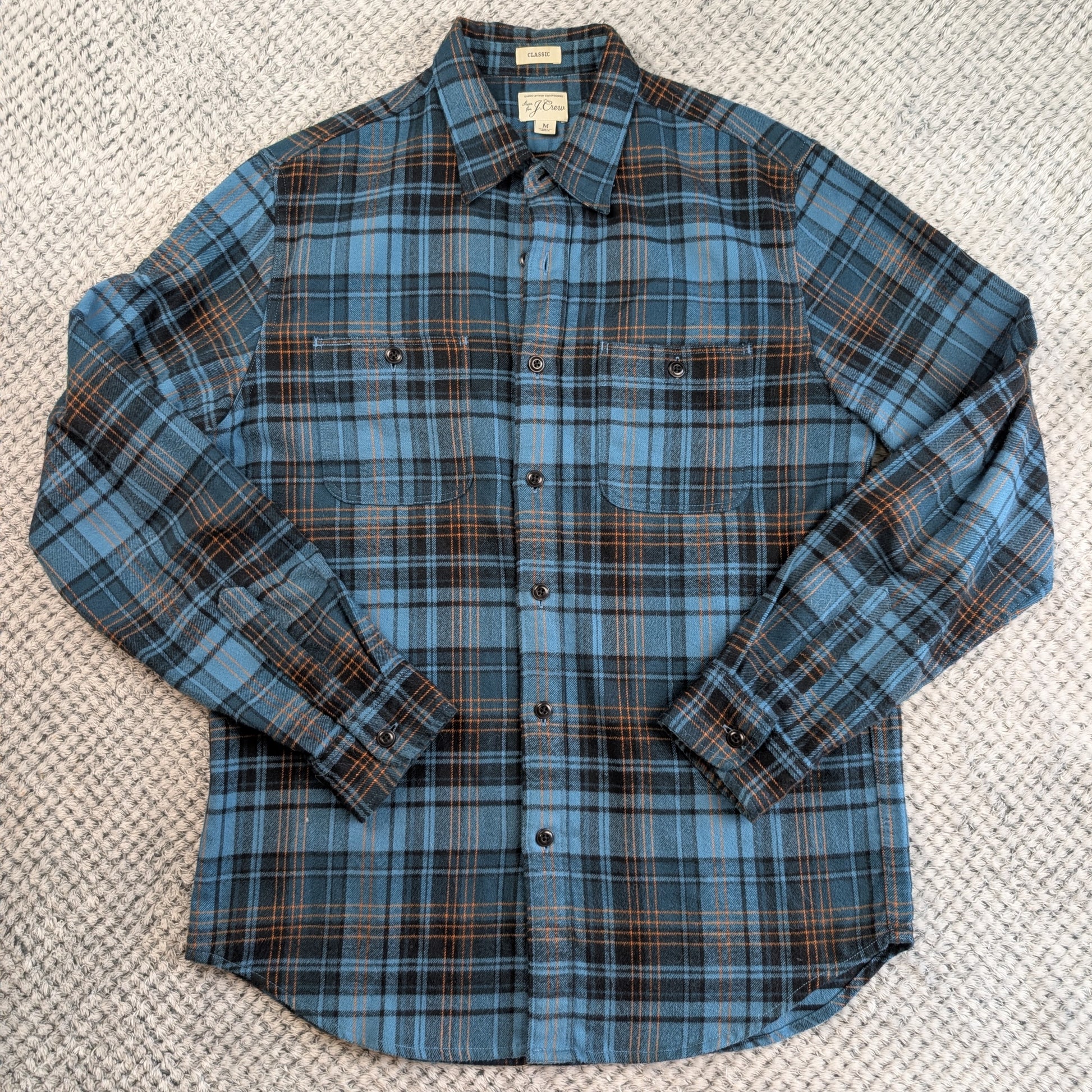 A long-sleeve button-up flannel shirt in a blue, black, and brown plaid pattern, laid flat on a textured gray woven rug.