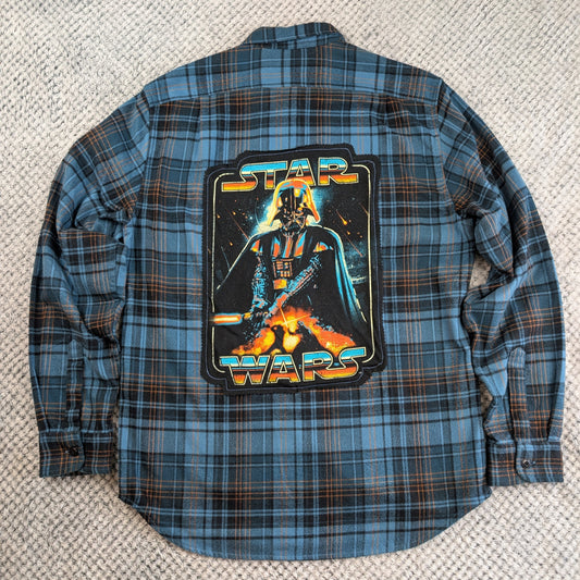 A blue and brown plaid Star Wars flannel shirt laid flat on a textured beige carpet, featuring a large graphic print of Darth Vader on the back with 'STAR WARS' text.