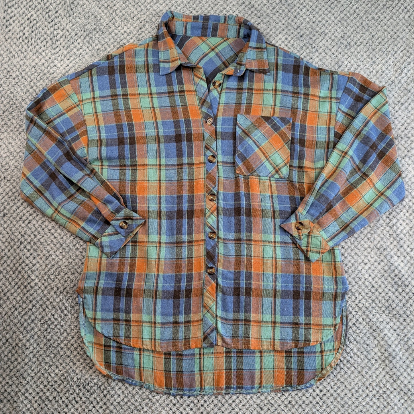 A long-sleeve plaid button-up shirt laid flat on a textured gray woven surface. The shirt features a multicolor plaid pattern with prominent orange, blue, green, and brown stripes.