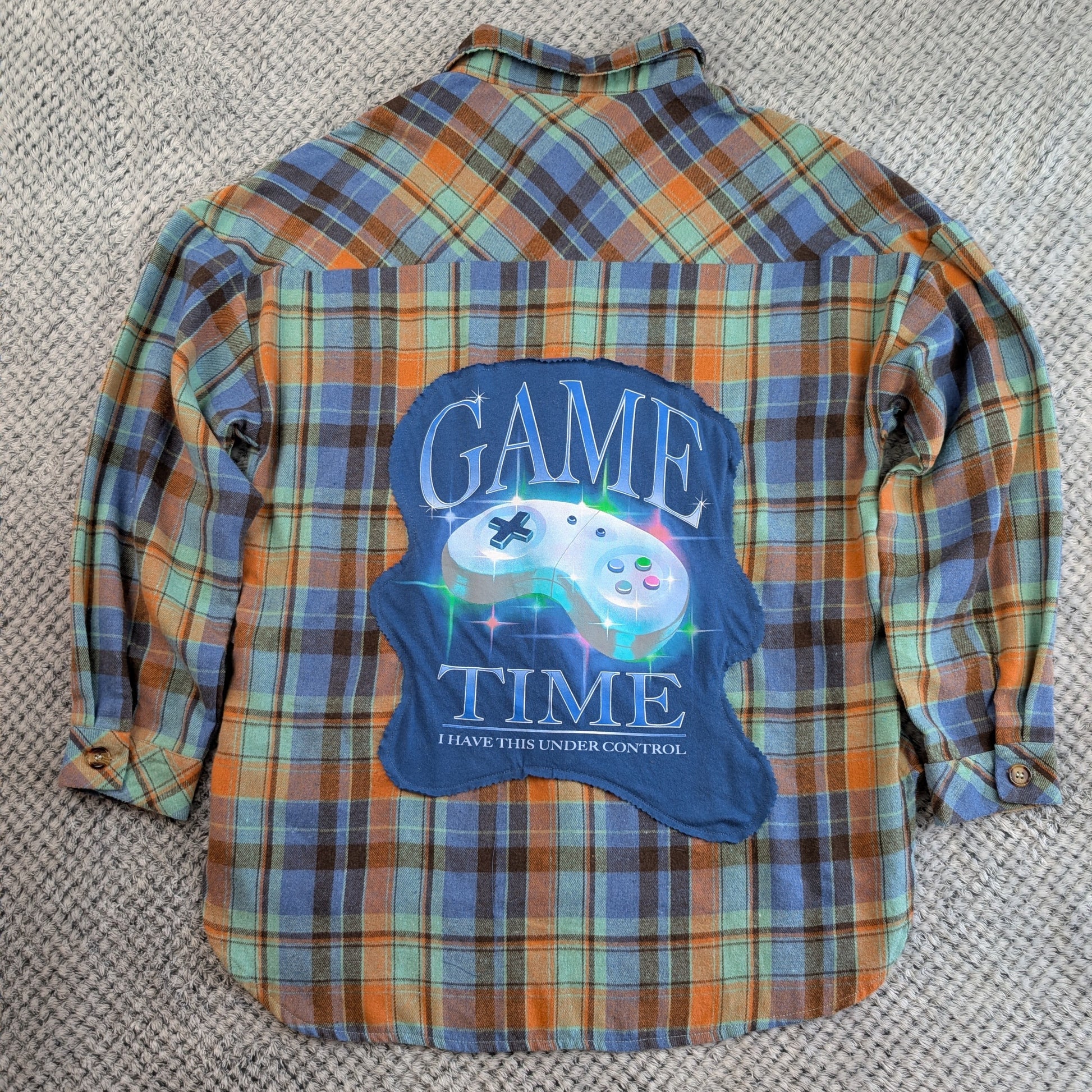 A long-sleeve plaid flannel shirt laid flat on a textured gray carpet. The shirt features a blue graphic patch on the back with a video game controller and the text 'GAME TIME' and 'I HAVE THIS UNDER CONTROL'.