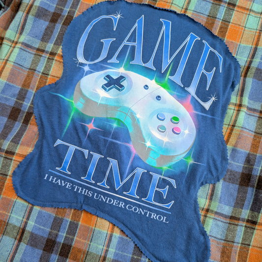 A blue fleece blanket with a 'GAME TIME I HAVE THIS UNDER CONTROL' graphic featuring a white game controller and colorful starburst effects, laid flat on a plaid blanket background.