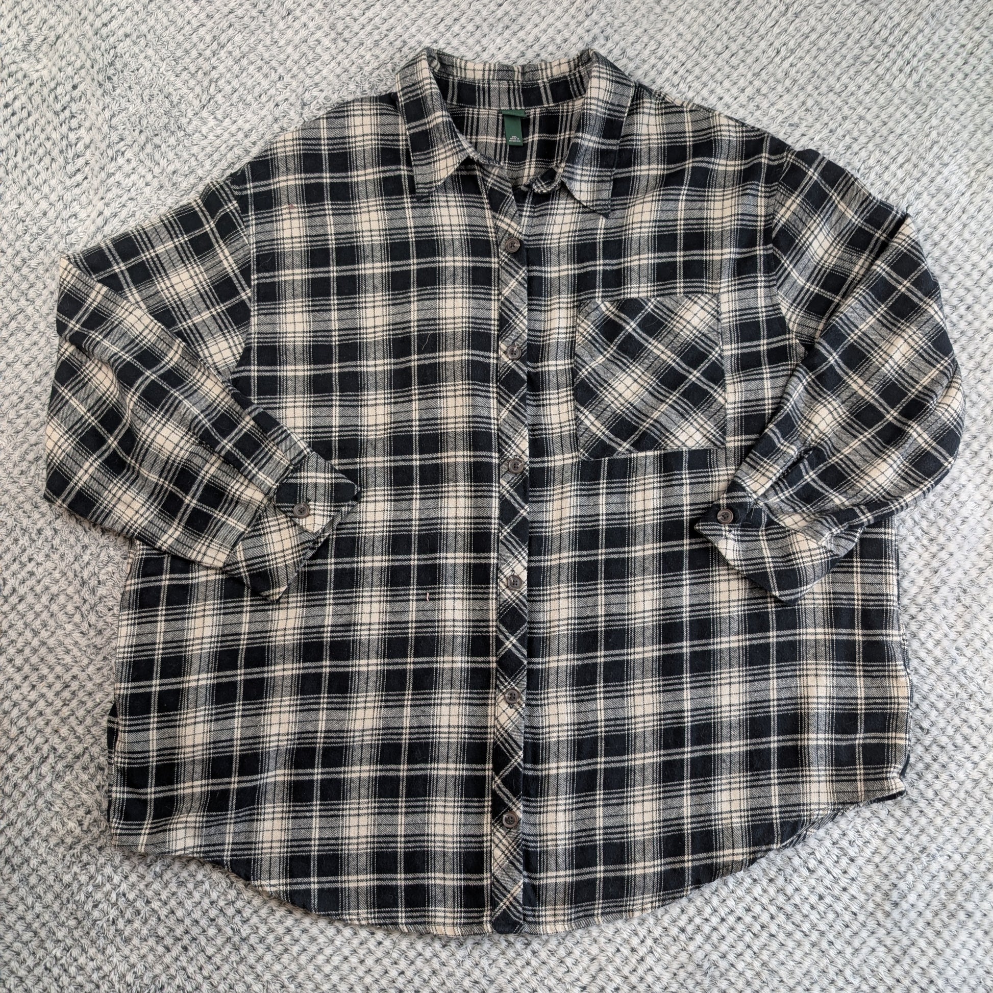A long-sleeve button-up flannel shirt with a black, white, and beige plaid pattern, laid flat on a textured gray carpet.
