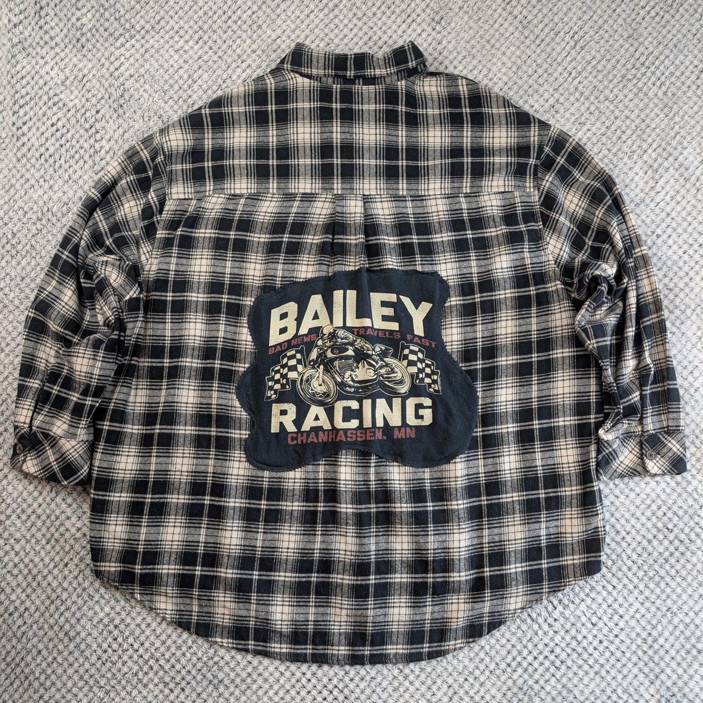 A vintage Bailey Racing plaid flannel shirt laid flat on a textured gray carpet. The shirt features a black, white, and beige plaid pattern with a large dark blue graphic patch on the back displaying 'BAILEY RACING' and a motorcycle illustration.