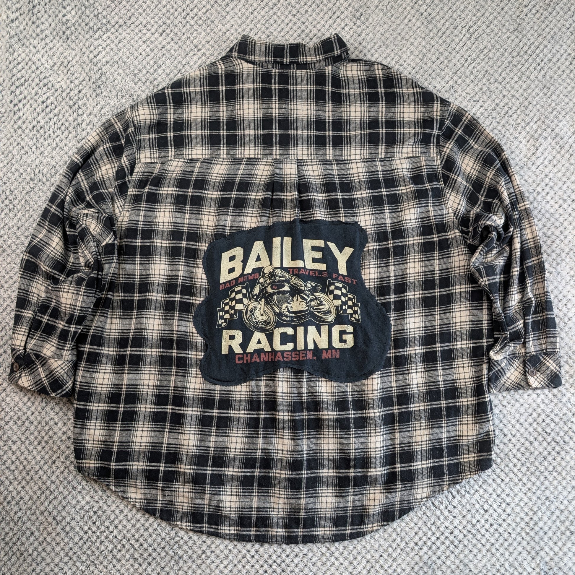 A vintage Bailey Racing plaid flannel shirt laid flat on a textured gray carpet. The shirt features a black, white, and beige plaid pattern with a large dark blue graphic patch on the back displaying 'BAILEY RACING' and a motorcycle illustration.
