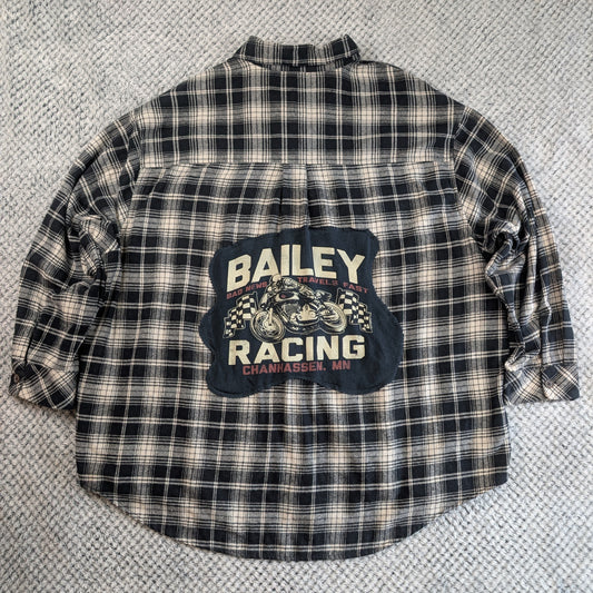 A vintage Bailey Racing plaid flannel shirt laid flat on a textured gray carpet. The shirt features a black, white, and beige plaid pattern with a large dark blue graphic patch on the back displaying 'BAILEY RACING' and a motorcycle illustration.